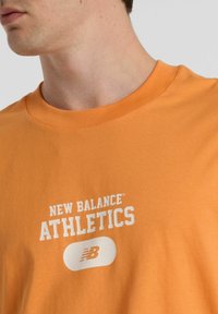Orange cotton t-shirt featuring "NEW BALANCE ATHLETICS" printed in white. Rounded neckline and soft texture, designed for comfort.
