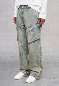 Pantalones cargo de denim azul claro, holgados, con múltiples bolsillos, costuras visibles y una textura deslavada. Combinados con zapatillas blancas.