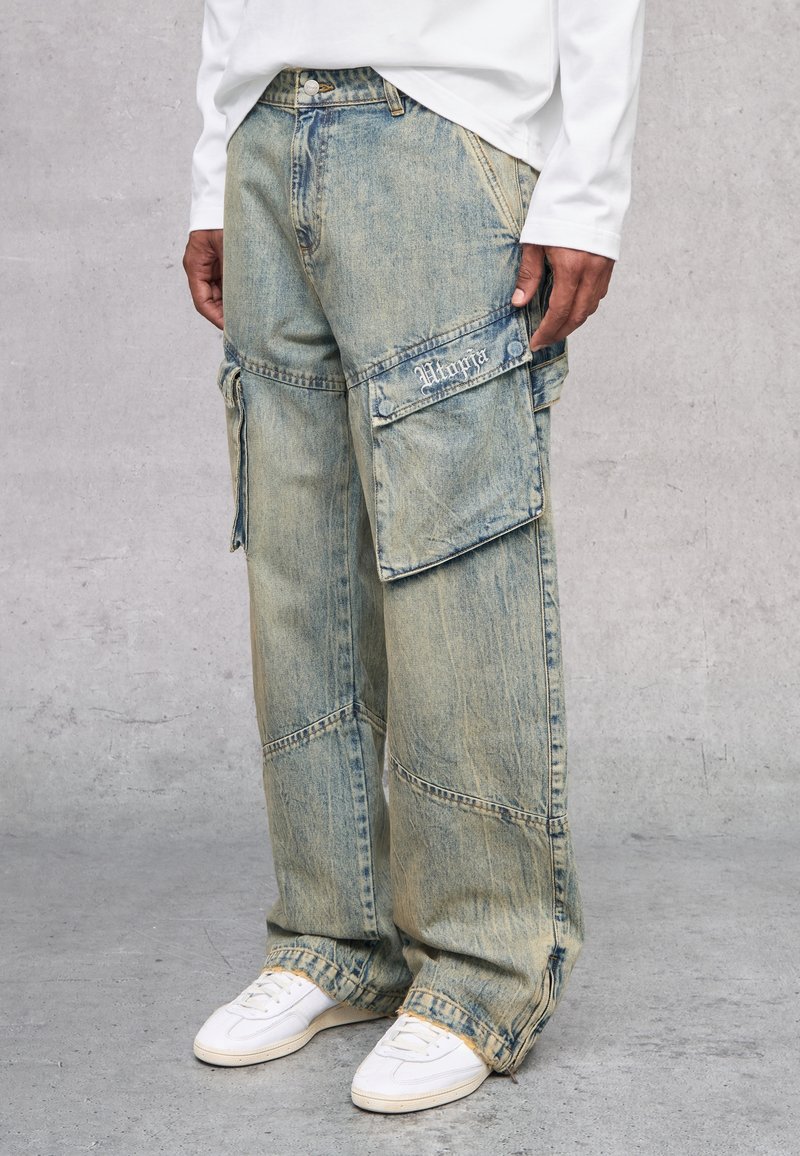 Pantalones cargo de denim azul claro, holgados, con múltiples bolsillos, costuras visibles y una textura deslavada. Combinados con zapatillas blancas.
