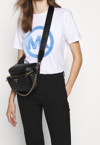 Black leather crossbody bag with a round shape, gold chain strap, and zippered compartment. Paired with a white t-shirt featuring a blue logo.