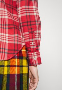 Röd rutig skjorta med knappar och vita och svarta accenter. Har rundad nederkant och en knapp i ärmslutet. Bärs med en flerfärgad rutig kjol.