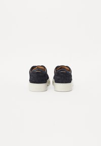 Mustat suede-tossut, joissa on kudottu rakenne, valkoinen kumipohja ja vaaleanruskea nahkavuori; takana on hienovarainen brändäys korkin päällä.