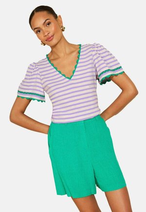 Kvinde iført en lilla og hvide stribet V-hals top med grønne bølgede kanter og grønne højtaljede shorts, stående med hænderne på hofterne.