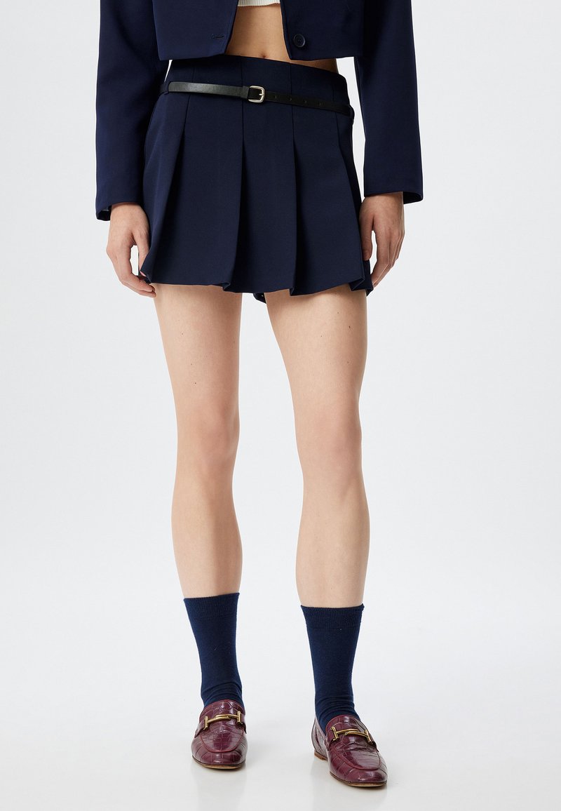 Tummansinsinen taitettu hame mustalla vyöllä, yhdistettynä tummansinsisiin sukkahousuihin ja viininpunaisiin loafereihin, joissa on kultaiset yksityiskohdat, ja esittää tyylikkään, muotoiltua designia.