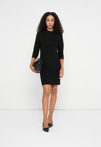 Donna con capelli ricci che indossa un vestito lungo da maglione nero, tiene una borsa nera ed è calzata con scarpe nere a punta, in piedi contro uno sfondo bianco.
