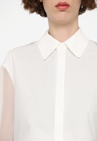 White collared shirt with opaque fabric and sheer sleeves. Features a button closure at the front and a smooth texture.