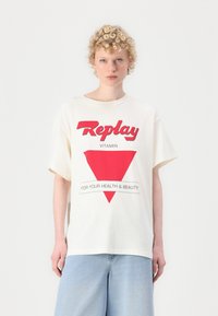 Biała, oversize'owa koszulka z krótkim rękawem z czerwonym nadrukiem graficznym, na którym widnieje napis "Replay Vitamin" oraz czerwony wzór w kształcie trójkąta. Koszulka ma okrągły dekolt i krótkie rękawy.