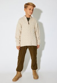 Cremefarbener, kabelgestrickter Pullover mit Halbzipper, olivgrüne Cargo-Hosen und braune Schnürboots. Der Pullover hat ribbelige Bündchen und einen Saum.