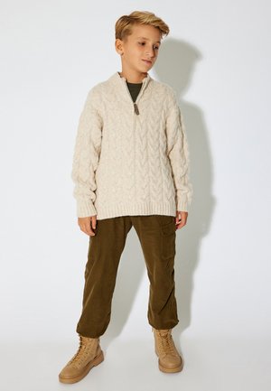 Suéter de punto trenzado color crema con media cremallera, pantalones cargo verdes y botas de cordones color beige. El suéter cuenta con puños y dobladillo acanalados.