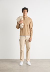 Camisa de polo de manga larga color marrón, pantalones beige y zapatillas blancas. El hombre lleva una chaqueta beige sobre el hombro. Diseño simple y casual.