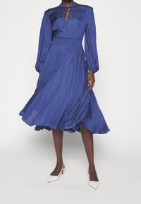 Vestido azul de manga larga con escote en forma de ojo de cerradura, tela plisada y cintura definida. Combinado con tacones blancos de punta afilada, que destacan un diseño fluido.