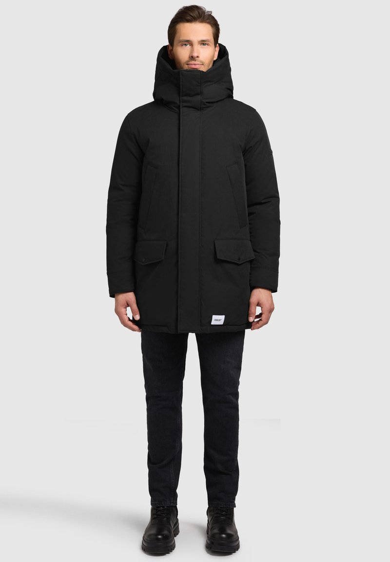 Czarna ocieplana parka z wysokim kołnierzem, dwiema przednimi kieszeniami i subtelnym logo. Tkanina ma gładką, matową fakturę.