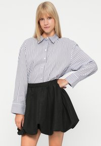 Jonge vrouw met blond haar draagt een losse, gestreepte blouse en een korte, zwarte klokrok, staande tegen een eenvoudige achtergrond.