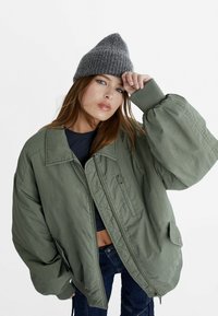 Jeune femme ajustant un bonnet tricoté gris, portant une veste oversize vert olive et un jean bleu foncé sur fond blanc.