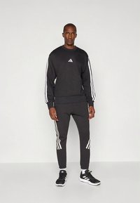 Μαύρο φούτερ με λευκό λογότυπο Adidas, σε συνδυασμό με μαύρο παντελόνι φόρμας με λευκές ρίγες στα πλαϊνά. Και τα δύο κατασκευασμένα από μαλακό ύφασμα.