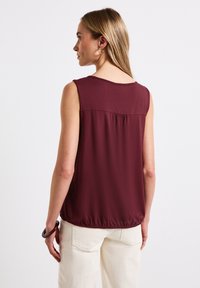 Top sin mangas en color burdeos con un corte relajado, detalle en la parte posterior con fruncido y dobladillo elástico. Se combina con pantalones de color claro que tienen una textura suave.