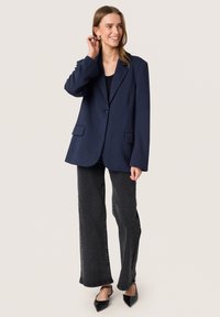 Marineblazer med en lang, afslappet pasform, to forlommer og enkeltknaplukning, parret med sorte vide jeans og spidse sko.