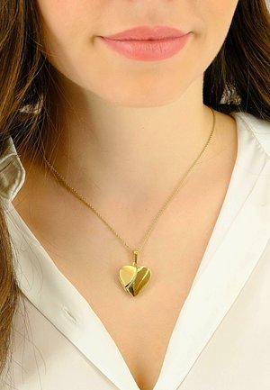 Pendentif en forme de cœur doré sur une fine chaîne, présentant un design à double finition avec une surface polie et une surface brossée, porté sur une blouse blanche.