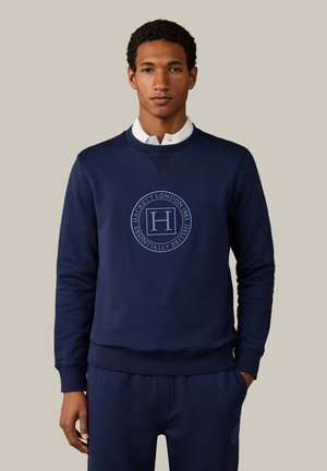 Jonge man draagt een marineblauwe trui en bijpassende broek met een rond Hackett London-logo, staand tegen een effen lichte achtergrond.