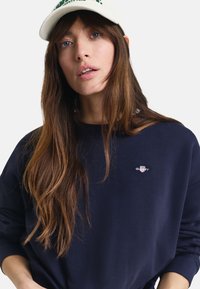 Marineblå sweatshirt med lange ermer, brodert logo på brystet, supplert med en kremfarget caps med grønn tekst. Glatt stofftekstur.