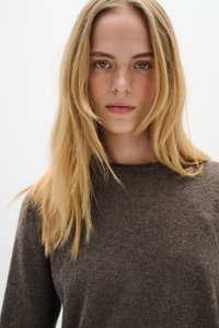Barna kötött pulóver kerek nyakkal, puha textúrával. A modellnek hosszú, egyenes, szőke haja van, és visszafogott sminkje.