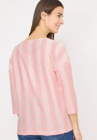 Kvinde med langt brunt hår iført en hvid og rød stribet langærmet skjorte og blå jeans, smilende og kiggede til siden.