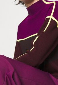 Gebreide trui met een hoge kraag, in paars en donkerbruin met gele accenten langs de naden en geometrische patronen. Gladde textuur.