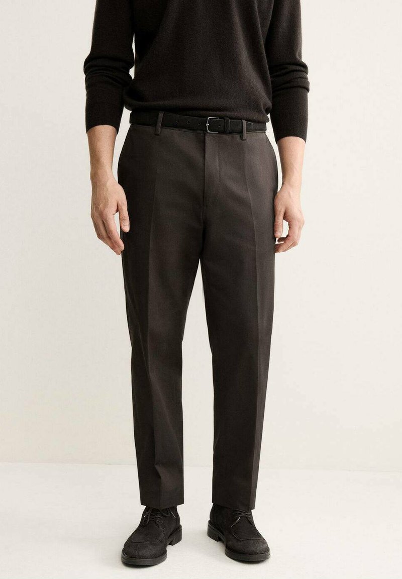 Pantalon gris foncé avec une coupe ajustée, présentant un devant plat et de jolis plis, associé à une ceinture noire et des chaussures en daim noires.