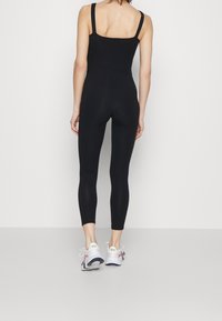 Kobieta stojąca tyłem, ubrana w czarny kombinezon bez rękawów i białe trampki Nike z kwiatowymi detalami.