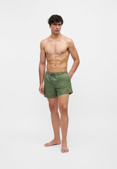 Homme torse nu aux cheveux foncés portant un short de bain vert à cordon, debout pieds nus sur un fond clair uni.