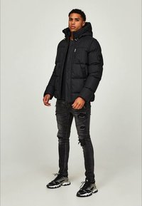 Giacca a vento nera con cappuccio, caratterizzata da una texture trapuntata e una finitura lucida. Abbinata a jeans skinny neri strappati e sneakers sportive.