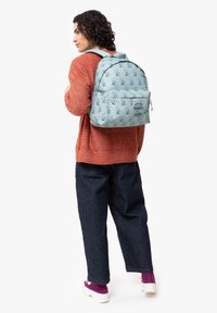 Ljusblå ryggsäck med ett upprepande mönster av tecknade karaktärer, framficka med dragkedja och vadderade remmar. Bärs över en rostfärgad tröja och mörka jeans.