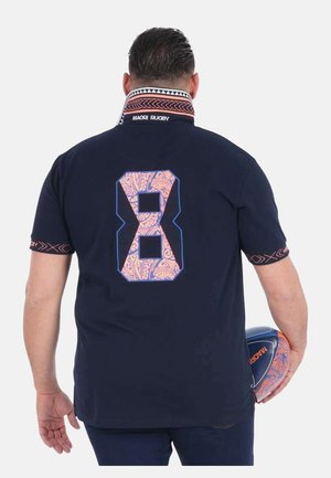 Homme tenant un ballon de rugby à motifs, portant un t-shirt bleu marine avec un grand chiffre 8 orné et des motifs maoris sur le col et les manches.