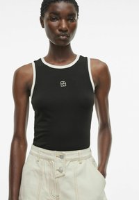 Zwarte mouwloze tanktop met crèmekleurige afwerking en een klein logo op de borst, gecombineerd met hoog-waisted lichtbeige broek met knoopdetails.