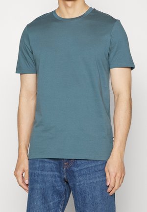 Homme portant un t-shirt bleu uni à col rond et manches courtes, et un jean bleu, debout les bras détendus le long du corps sur un fond blanc.