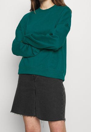 Kvinde iført en mørkegrøn sweatshirt med lange ærmer og en sort denimnederdel med frynser, armene krydsede, stående op ad en ensfarvet væg.