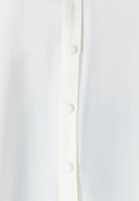 Detail eines weißen Hemd-Oberteils aus glattem Stoff, mit fünf gleichmäßig verteilten Knöpfen und einer weichen Textur entlang der Knopfleiste.