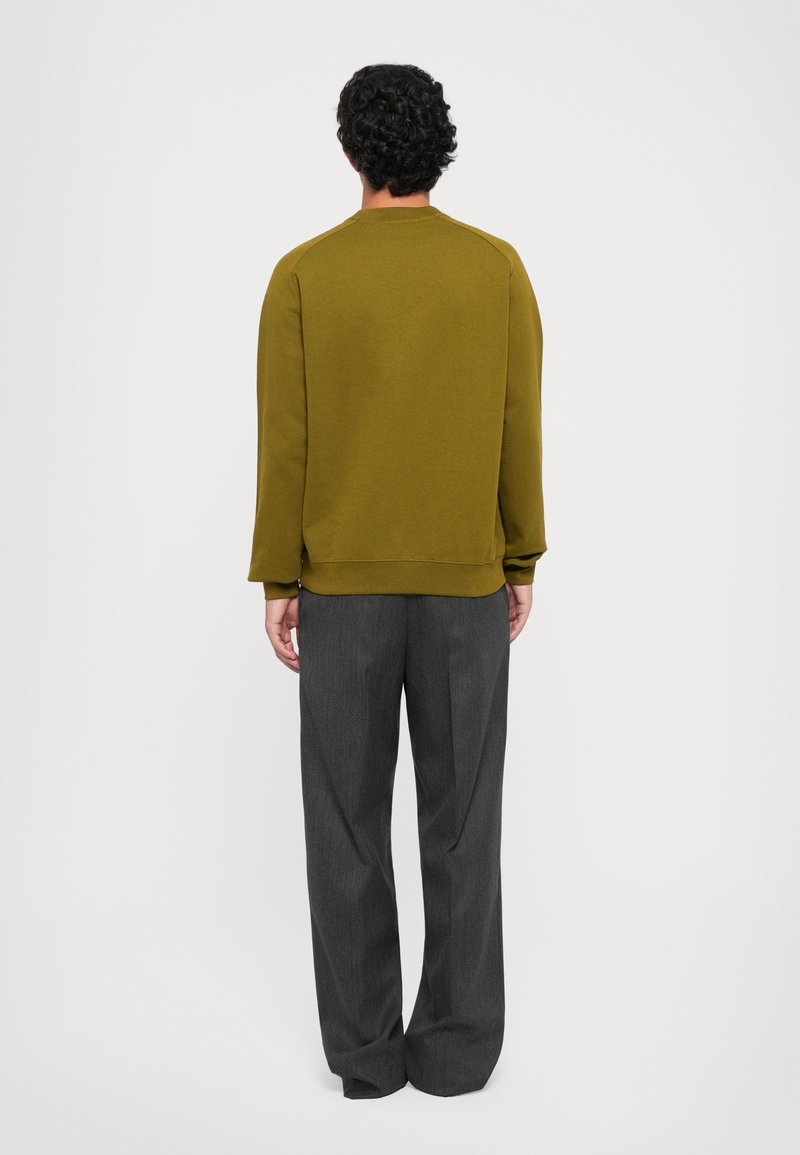 Homme aux cheveux noirs bouclés tourné vers le dos, portant un pull vert olive et un pantalon gris foncé ample, sur un fond blanc uni.