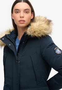 Parka bleu marine avec capuche à fourrure, fermeture éclair et poches à rabat. Présente un écusson sur la manche et une texture lisse.