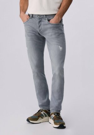 Uomo che indossa jeans aderenti grigio chiaro con dettagli leggermente usurati e sneakers multicolore, con le mani infilate nelle tasche.