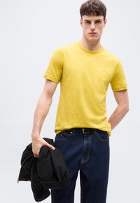 Νέος άνδρας που φοράει κίτρινο κοντομάνικο πουκάμισο και σκούρο τζιν κρατάει ένα μαύρο σακάκι στο αριστερό του χέρι, μπροστά σε ένα απλό φόντο.