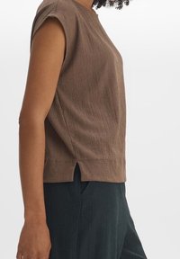 Sido vy av en person klädd i en strukturerad brun ärmlös topp med en liten sid slits och mörka strukturerade byxor.