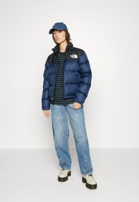Marineblå pufferjakke med sorte skuldre, hvitt logo, stripete langermet skjorte, lyseblå jeans og beige platåsko.
