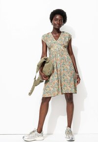Robe en coton floral avec un décolleté en V, des manches courtes et une taille froncée. Sac olive tenu dans une main, chaussures de sport argentées aux pieds.