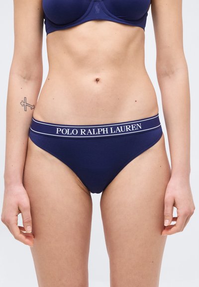 Γυναίκα που φορά σετ εσωρούχων Polo Ralph Lauren σε ναυτικό μπλε, με εμφανές τον κορμό και τα χέρια της και ένα μικρό τατουάζ σταυρού στο αριστερό αντιβράχιο.