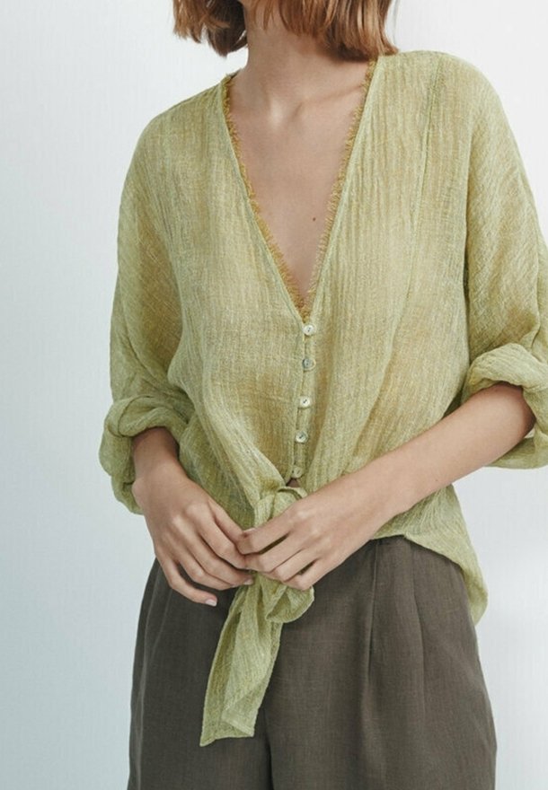 Personne portant un chemisier texturé vert clair avec manches retroussées et boutons sur le devant noués à la taille, associé à un pantalon sombre et ample.