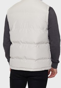 Wattierte Weste in Hellgrau mit gestepptem Design, Rundhalsausschnitt und glatter Textur, getragen über einem dunklen Langarmshirt.