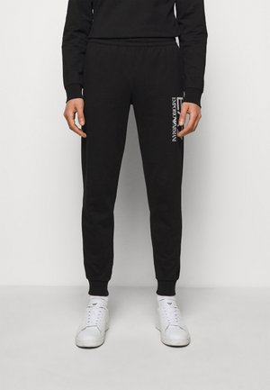 Personne portant un jogger noir slim avec le logo blanc Emporio Armani sur la cuisse gauche, associé à des baskets blanches et un haut noir assorti.
