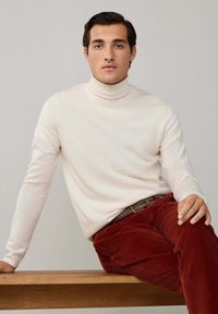 Cremfarbener Rollkragenpullover aus weichem Strickstoff, kombiniert mit rostrotfarbener Cordhose. Das Model sitzt auf einer Holzbank und präsentiert die Passform des Outfits.
