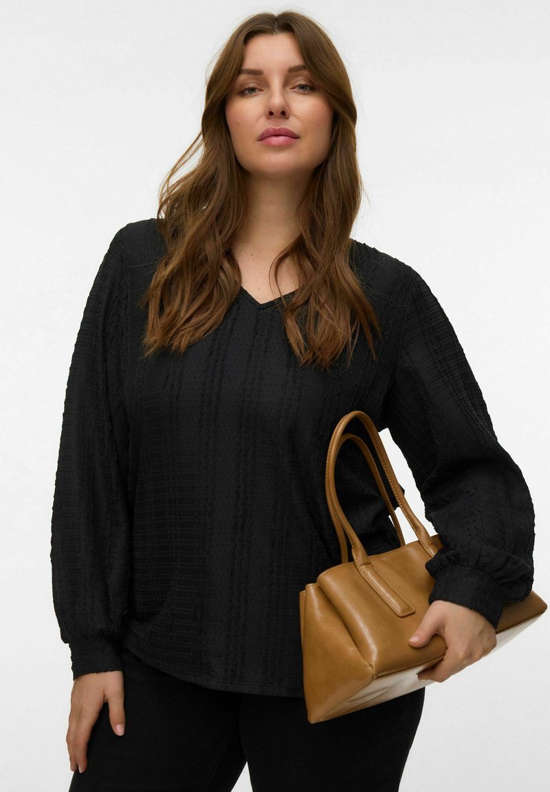 Haut noir texturé avec un col en V et des manches longues, associé à un sac à main marron structuré. La tenue présente une coupe décontractée.